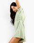 A woman modeling a light sage green bohemian-style mini dress with intricate eyelet embroidery on the chest, posed against a clean white background.