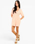 A woman modeling a light peach short-sleeve romper dress with a lace V-neck bodice, paired with beige platform sandals, against a plain white studio background.