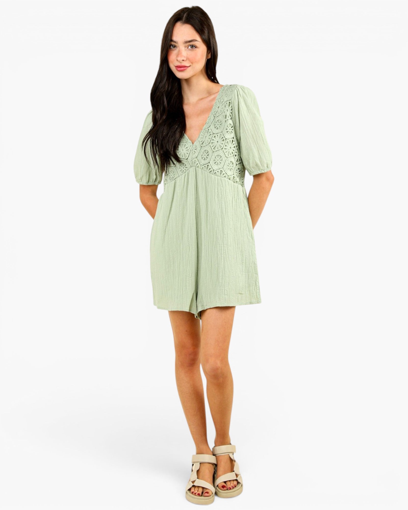 A woman modeling a light sage green romper with short puff sleeves, a V-neckline, and a floral lace yoke detail, paired with beige espadrille-style sandals against a plain white studio background.