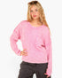 A woman modeling a pink fuzzy knit crewneck sweater with a front chest pocket, paired with dark gray jeans, against a plain white background.