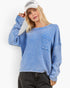 A woman wearing a light blue textured knit crewneck sweater with a chest pocket, paired with black jeans and a white baseball cap, against a plain white background.