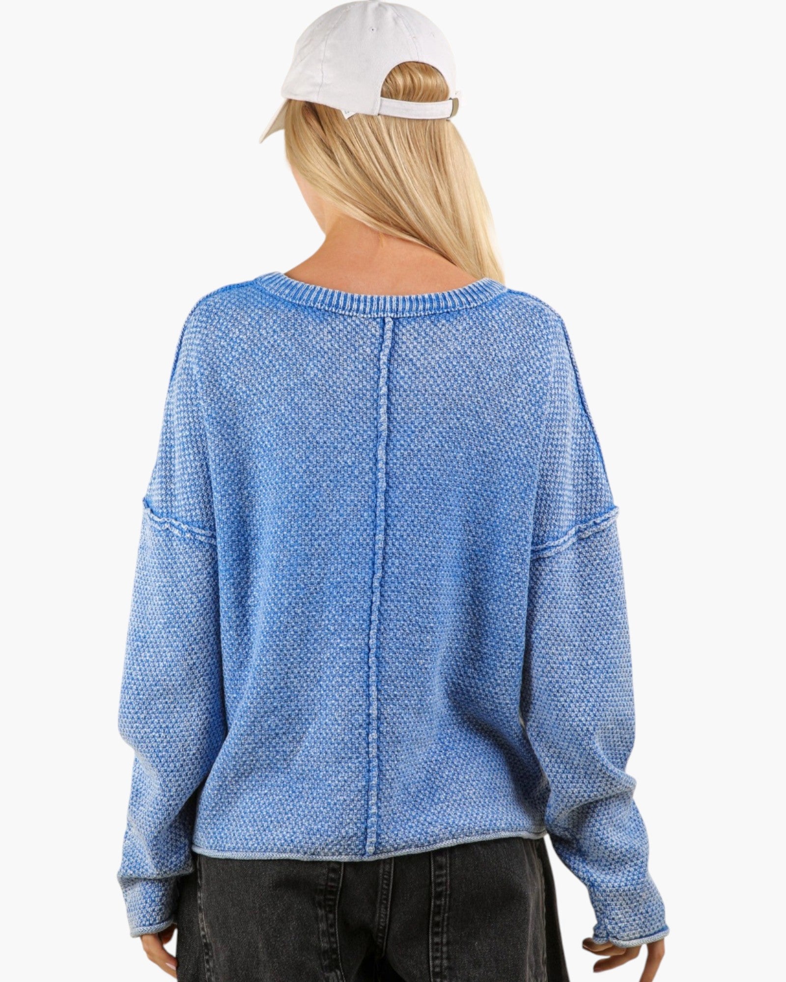 A woman viewed from the back wearing a light blue textured knit sweater, black jeans, and a white baseball cap against a plain white background.