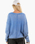 A woman viewed from the back wearing a light blue textured knit sweater, black jeans, and a white baseball cap against a plain white background.
