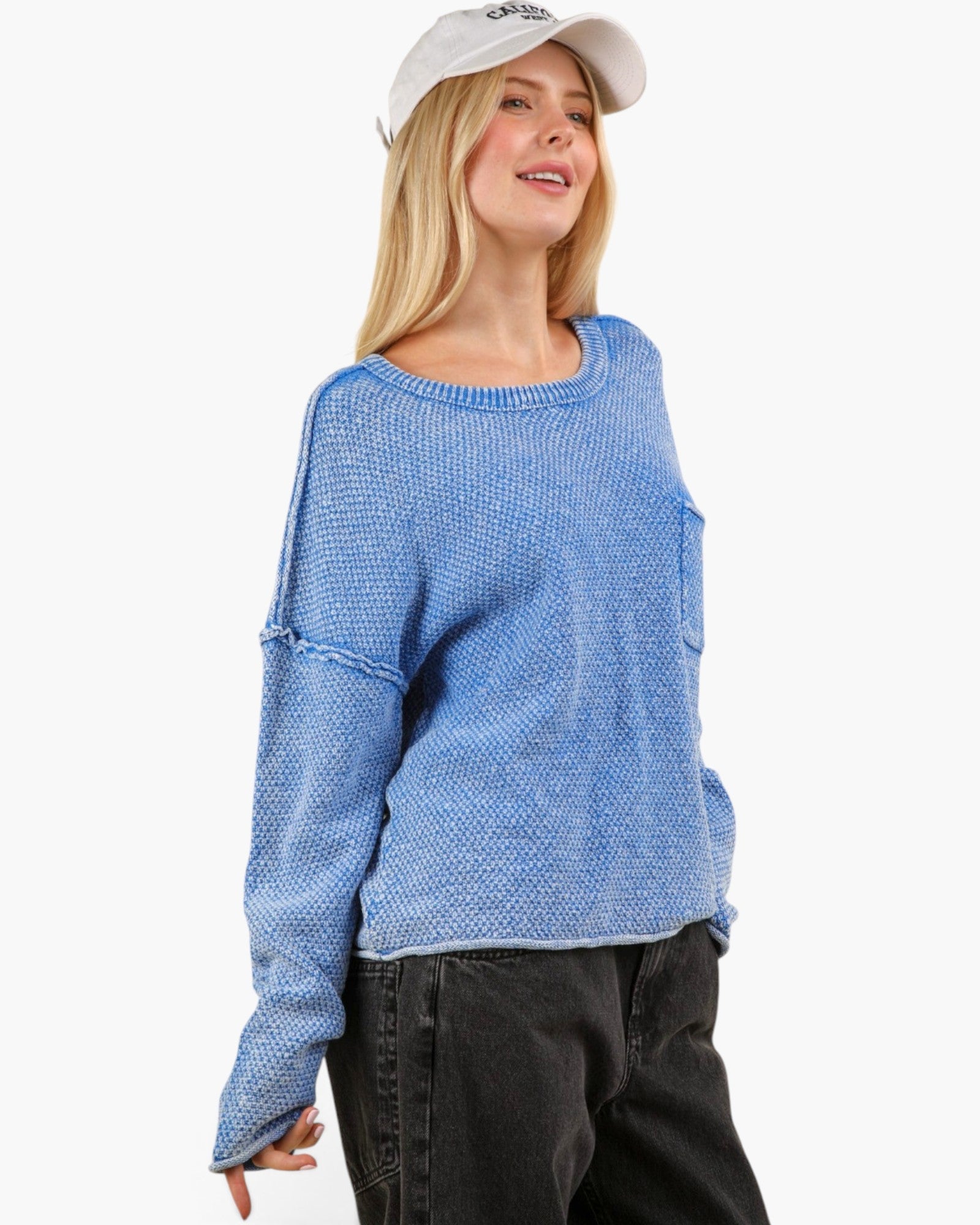 A woman wearing a light blue textured knit sweater with distressed details on the sleeves, paired with black jeans and a white baseball cap, set against a plain white background.
