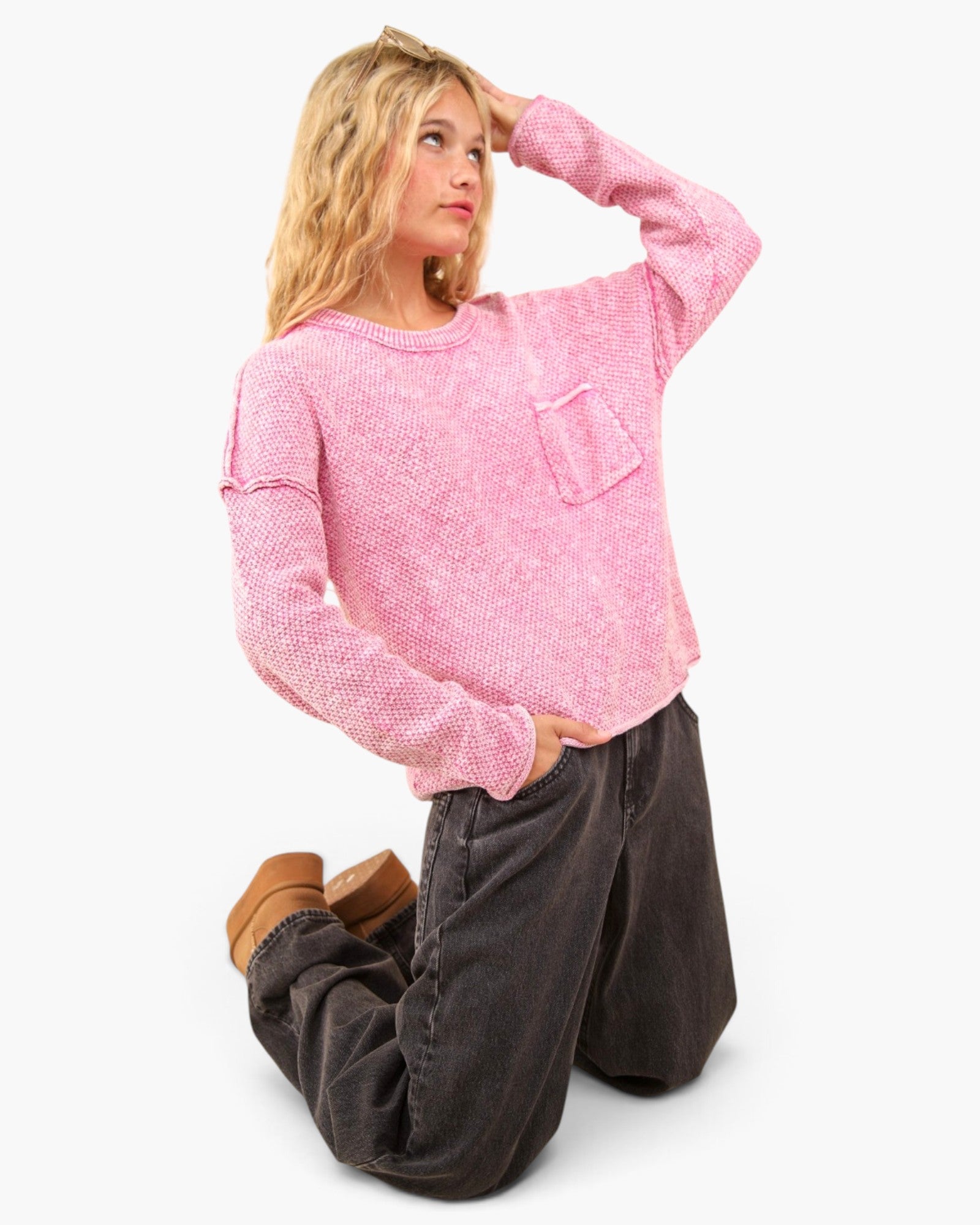 A woman kneeling on a light gray studio background wearing a pink textured knit sweater with a chest pocket, dark gray wide-leg pants, and brown boots.