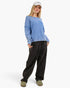 A woman modeling a light blue off-the-shoulder knit sweater, black wide-leg sweatpants, and olive green slip-on shoes against a plain white studio background.
