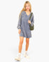 A young woman modeling a long-sleeve, V-neck, oversized heathered blue-gray knit dress. She is carrying an olive green shoulder bag and wearing off-white lace-up boots against a solid white studio background.