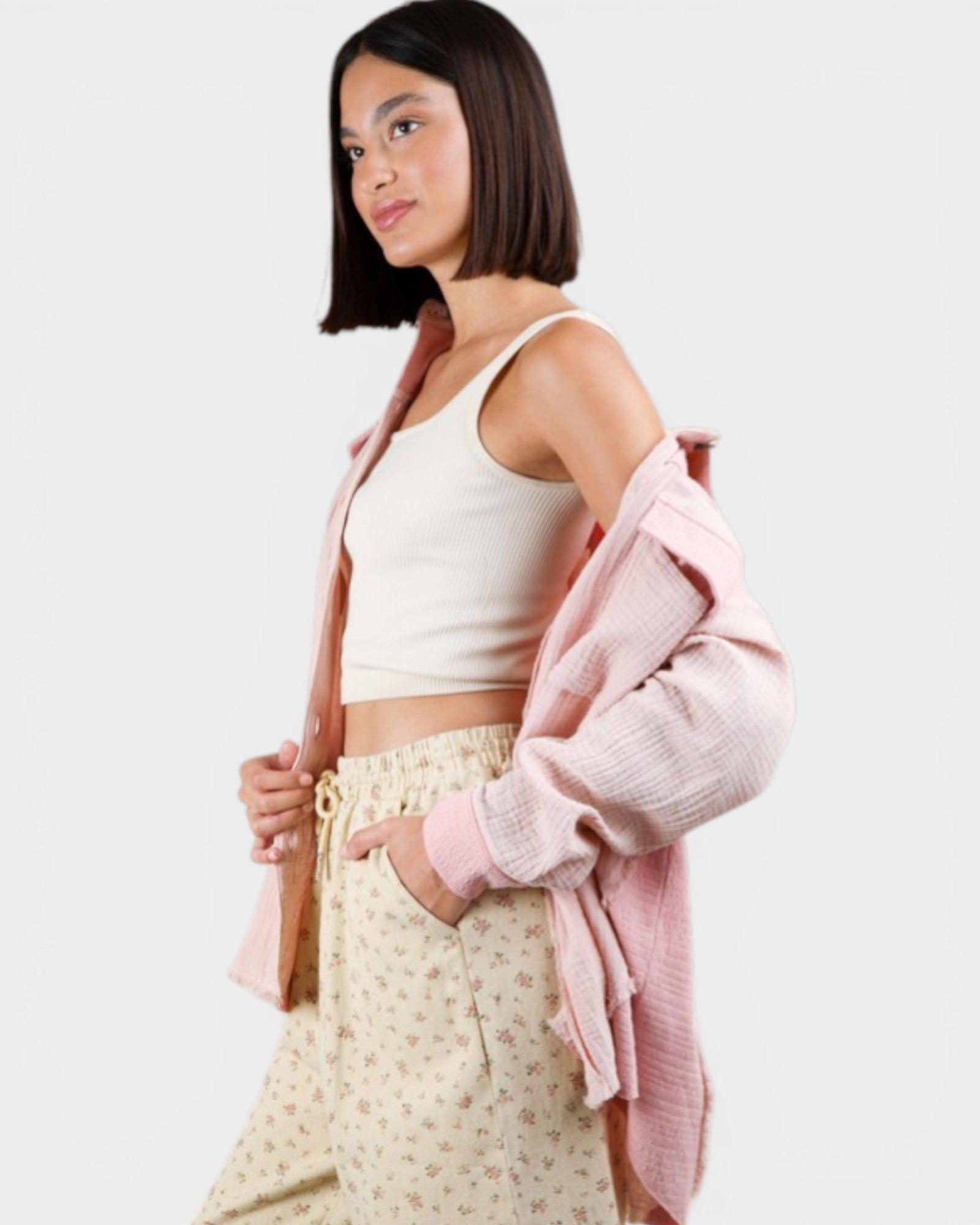 Woman wearing a white tank top and light-colored pants with a pink cardigan on a plain background