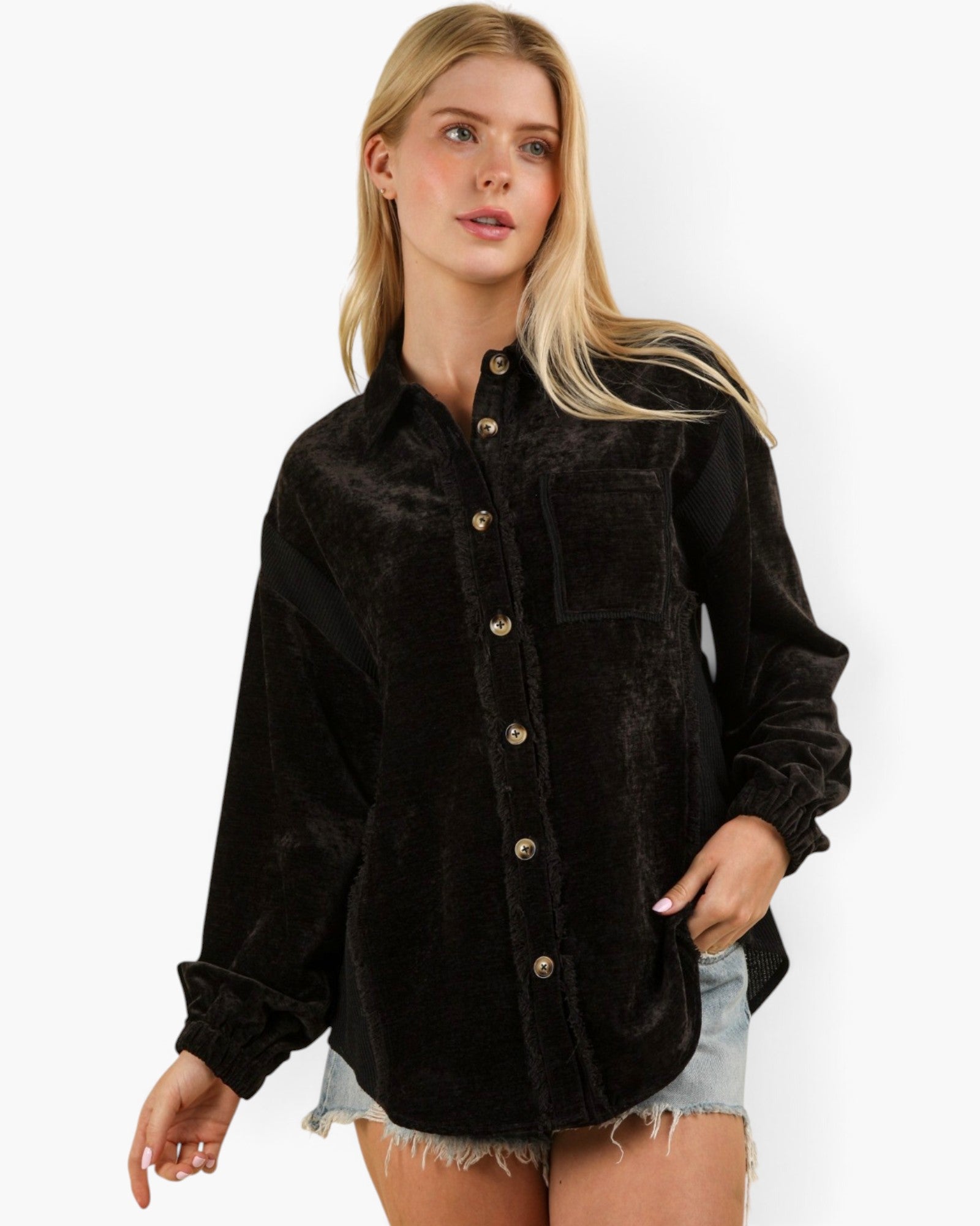 A woman modeling a black long-sleeve button-front shirt jacket made of textured corduroy or velvety fabric, paired with light denim shorts, against a plain white studio background.