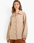 A woman wearing a beige button-up shirt jacket with textured fringe trim and elastic cuffs, paired with brown pants, against a plain white studio background.
