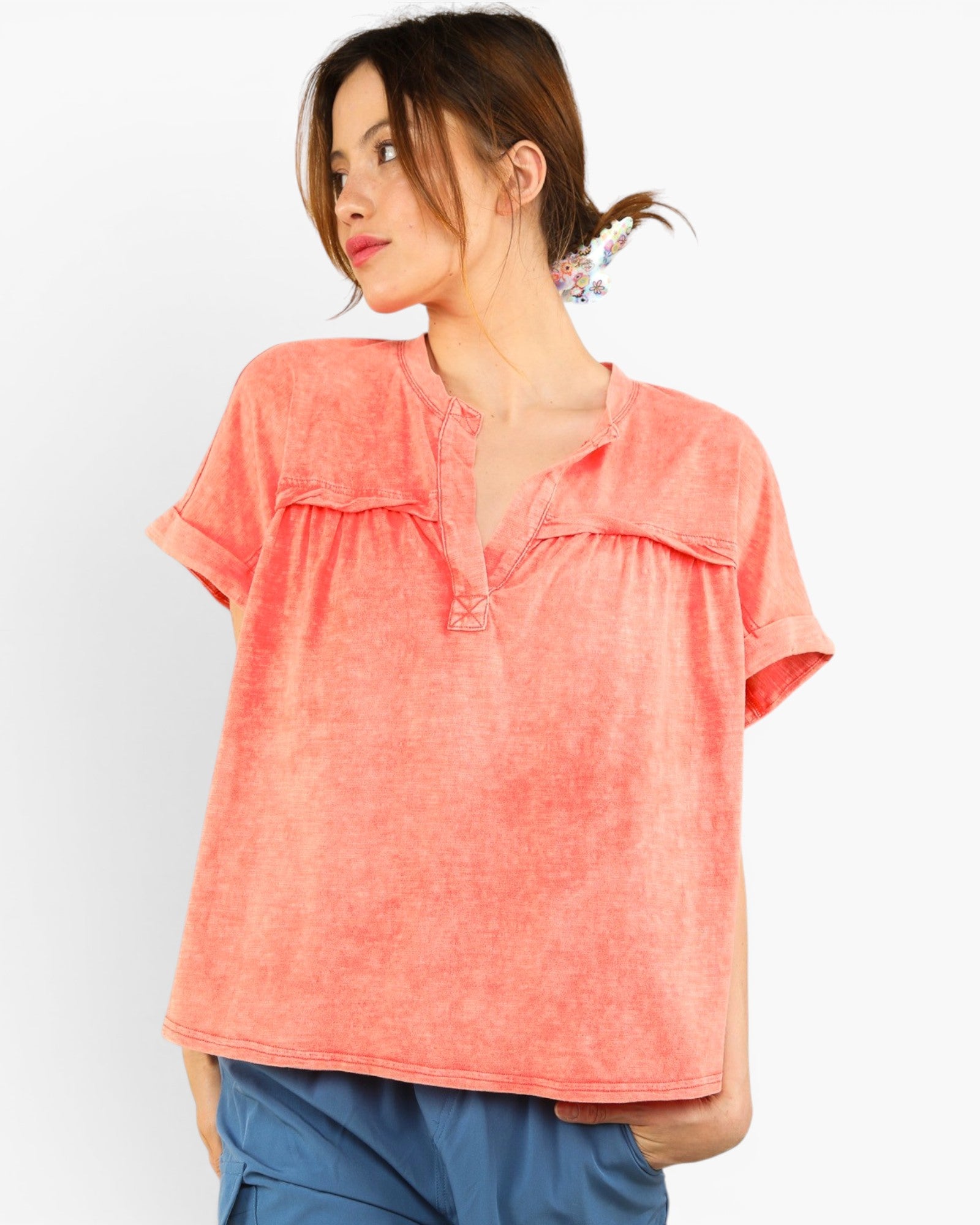 A woman wearing a short-sleeve coral pink top with a split neckline and gathered front detail, paired with blue pants, against a plain light background.