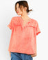 A woman wearing a short-sleeve coral pink top with a split neckline and gathered front detail, paired with blue pants, against a plain light background.
