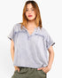 A woman wearing a light gray heathered short-sleeve top with a split V-neck and ruffle details, paired with olive green pants, against a plain white background.