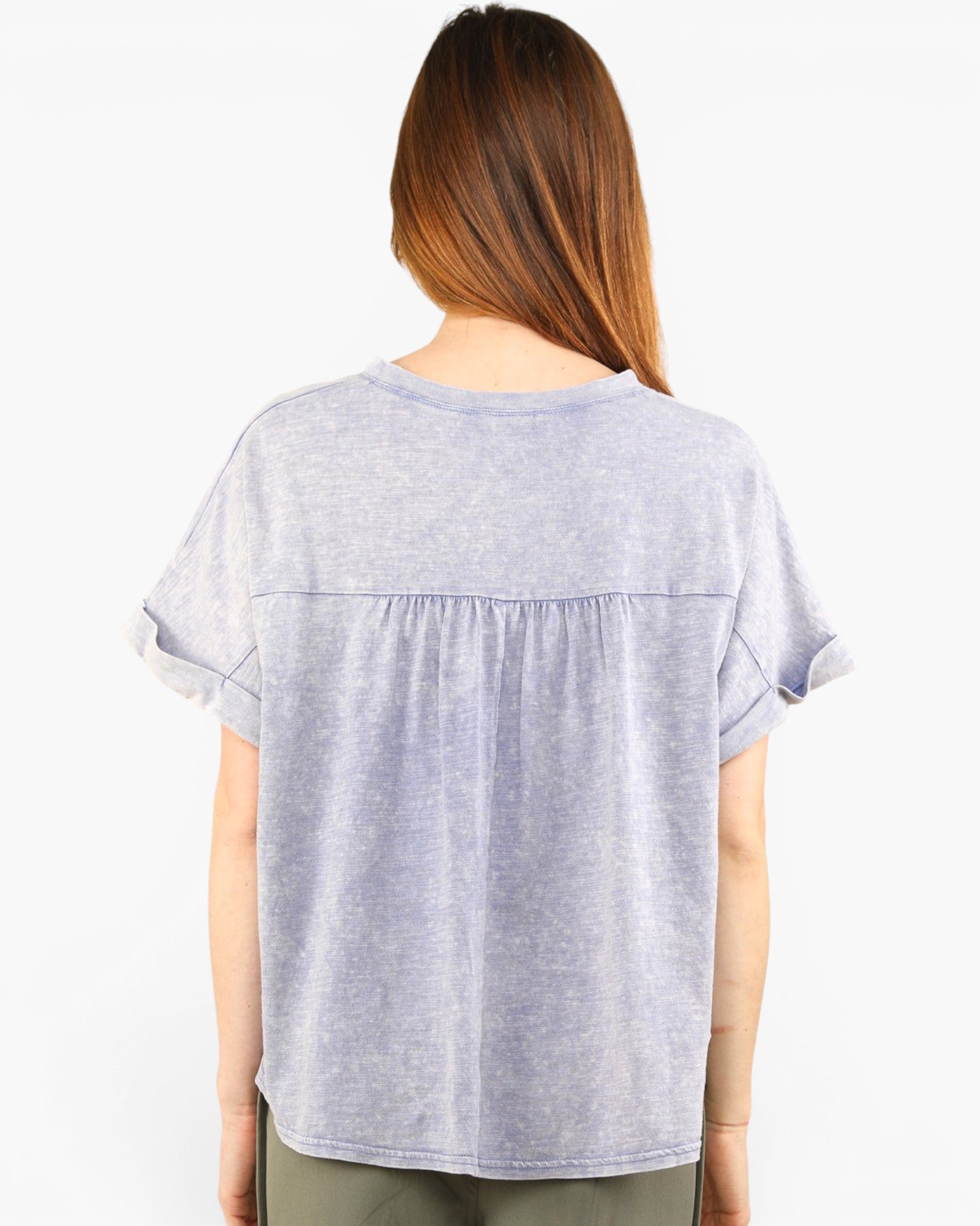 A woman viewed from the back wearing a light heather gray short-sleeve top with a gathered seam across the upper back and ruffled sleeve hems, paired with olive green pants, against a plain white background.