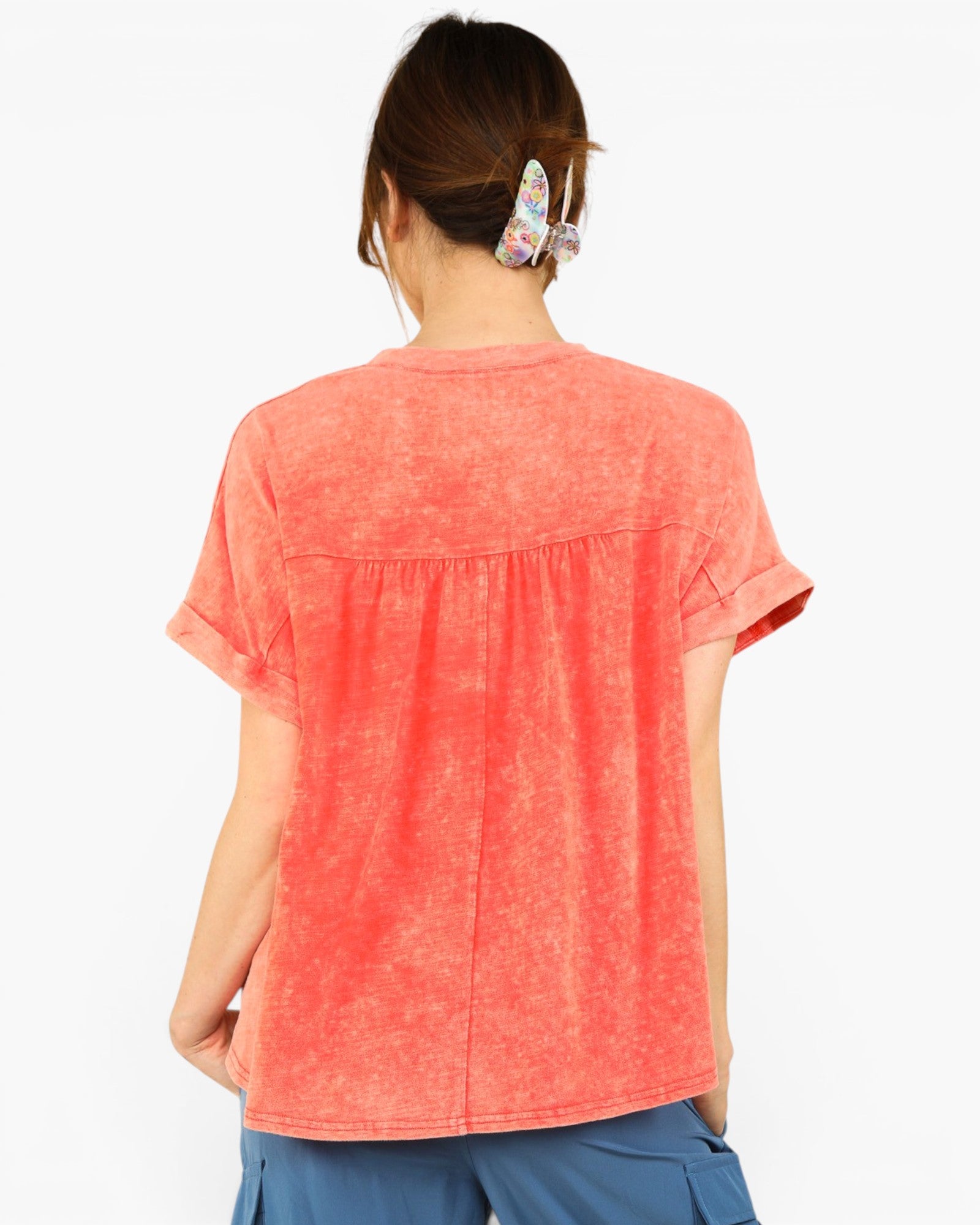 A woman viewed from the back wearing a short-sleeve coral orange top with a gathered waist seam and blue pants, against a plain white background.