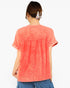 A woman viewed from the back wearing a short-sleeve coral orange top with a gathered waist seam and blue pants, against a plain white background.