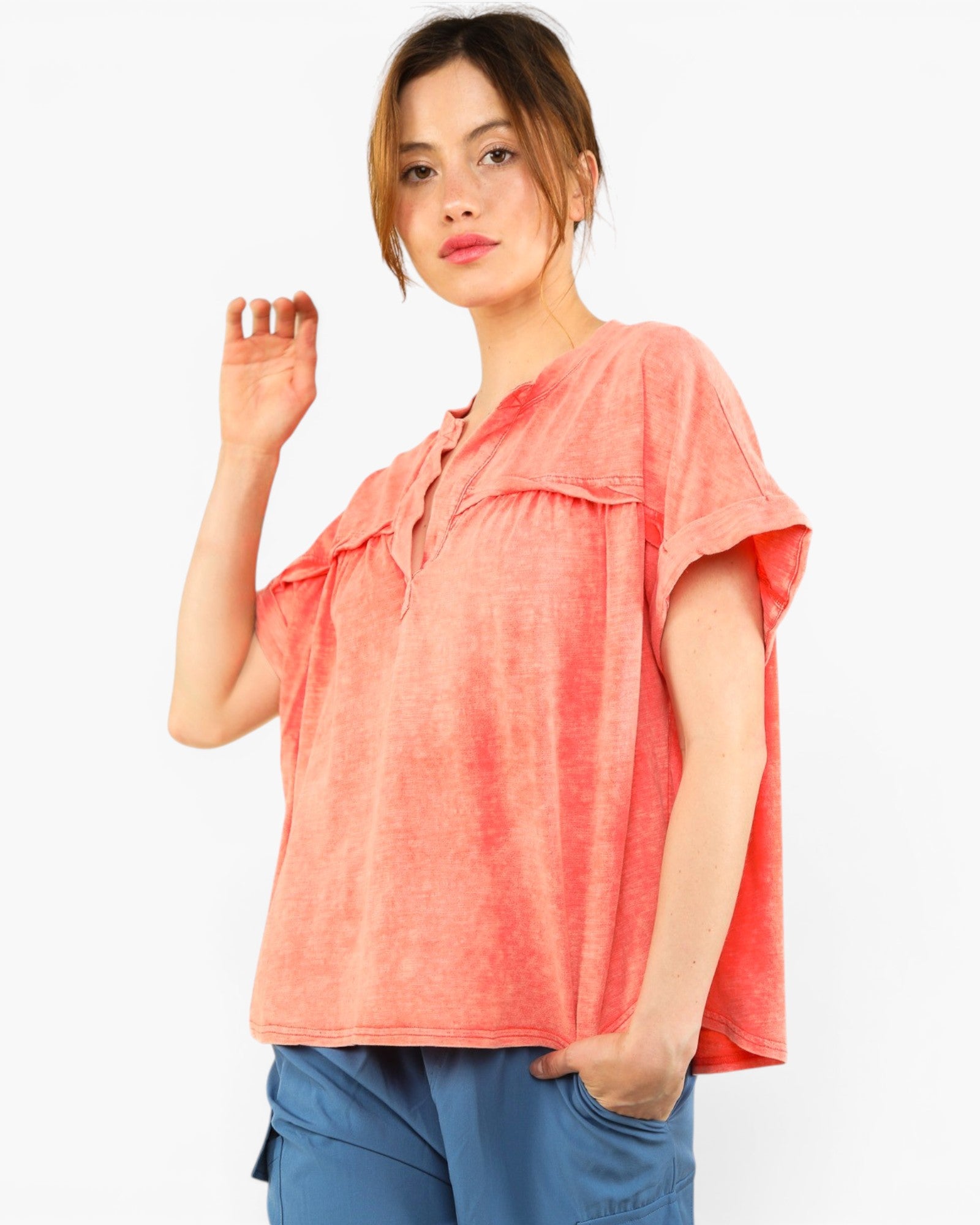 A woman modeling a short-sleeve coral pink top with a split neckline and ruffle details, paired with blue pants, against a plain white background.