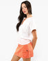 A woman wearing a white short-sleeve top and a coral orange denim mini skirt with a front flap pocket, photographed against a plain light gray studio background.