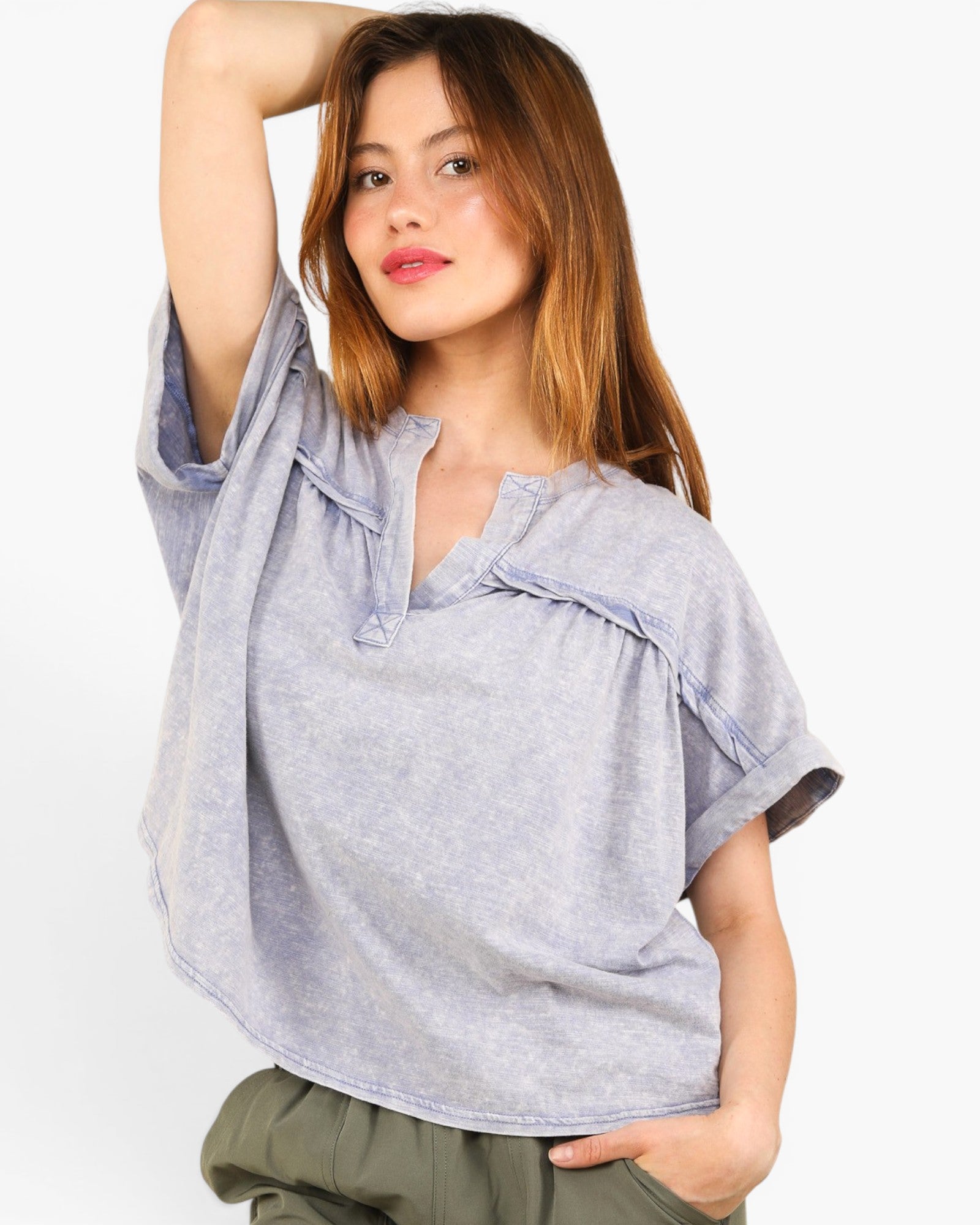 A woman wearing a light gray heathered short-sleeve top with ruffle trim and a split neckline, paired with olive green pants, against a plain white background.