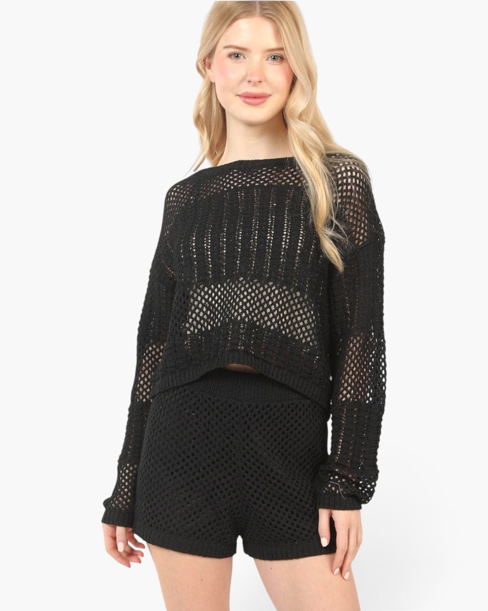 A blonde woman modeling a black long-sleeve knit crop top with a sheer openwork pattern, paired with matching black shorts, against a plain white studio background.