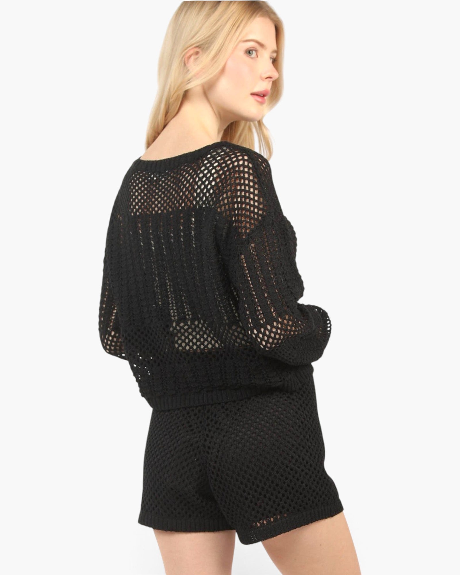 A woman wearing a black open-knit long-sleeve sweater and matching black shorts, shown from a side-back angle against a plain light gray studio background.
