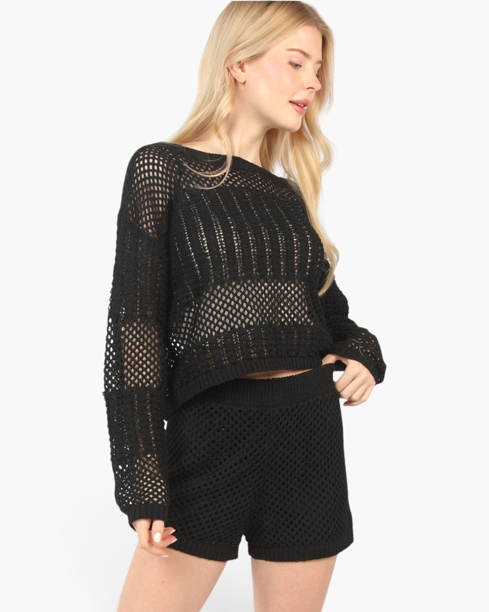 A blonde model wearing a black open-knit cropped sweater and matching black knit shorts against a plain white background.