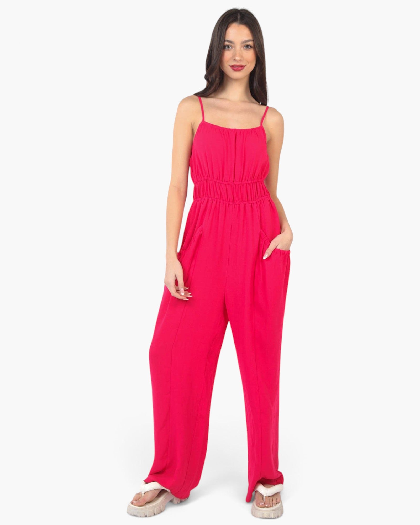 A woman modeling a bright pink sleeveless jumpsuit with thin spaghetti straps, a smocked waist, and wide-leg pants, standing against a plain white studio background.
