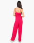 A woman viewed from the back wearing a bright pink sleeveless jumpsuit with thin straps, a gathered smocked bodice, and wide legs, against a plain light background.
