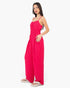 A woman modeling a bright pink sleeveless jumpsuit with thin straps, a gathered waist, and wide-leg pants, paired with light-colored sandals against a plain white background.