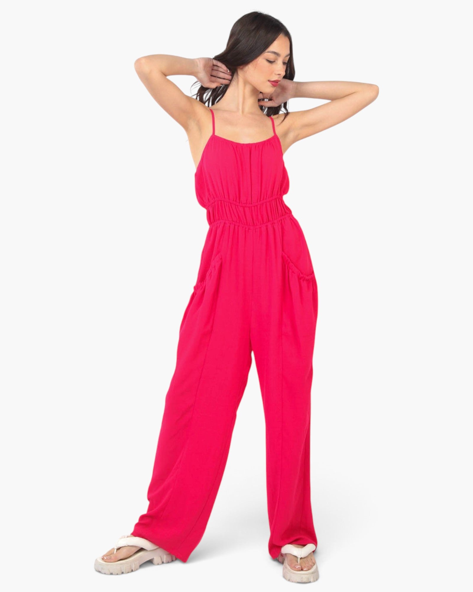 A woman modeling a bright pink sleeveless jumpsuit with thin straps, a gathered waist, and wide legs, paired with white slide sandals, against a plain light gray background.