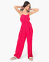 A woman modeling a bright pink sleeveless jumpsuit with thin straps, a gathered waist, and wide legs, paired with white slide sandals, against a plain light gray background.