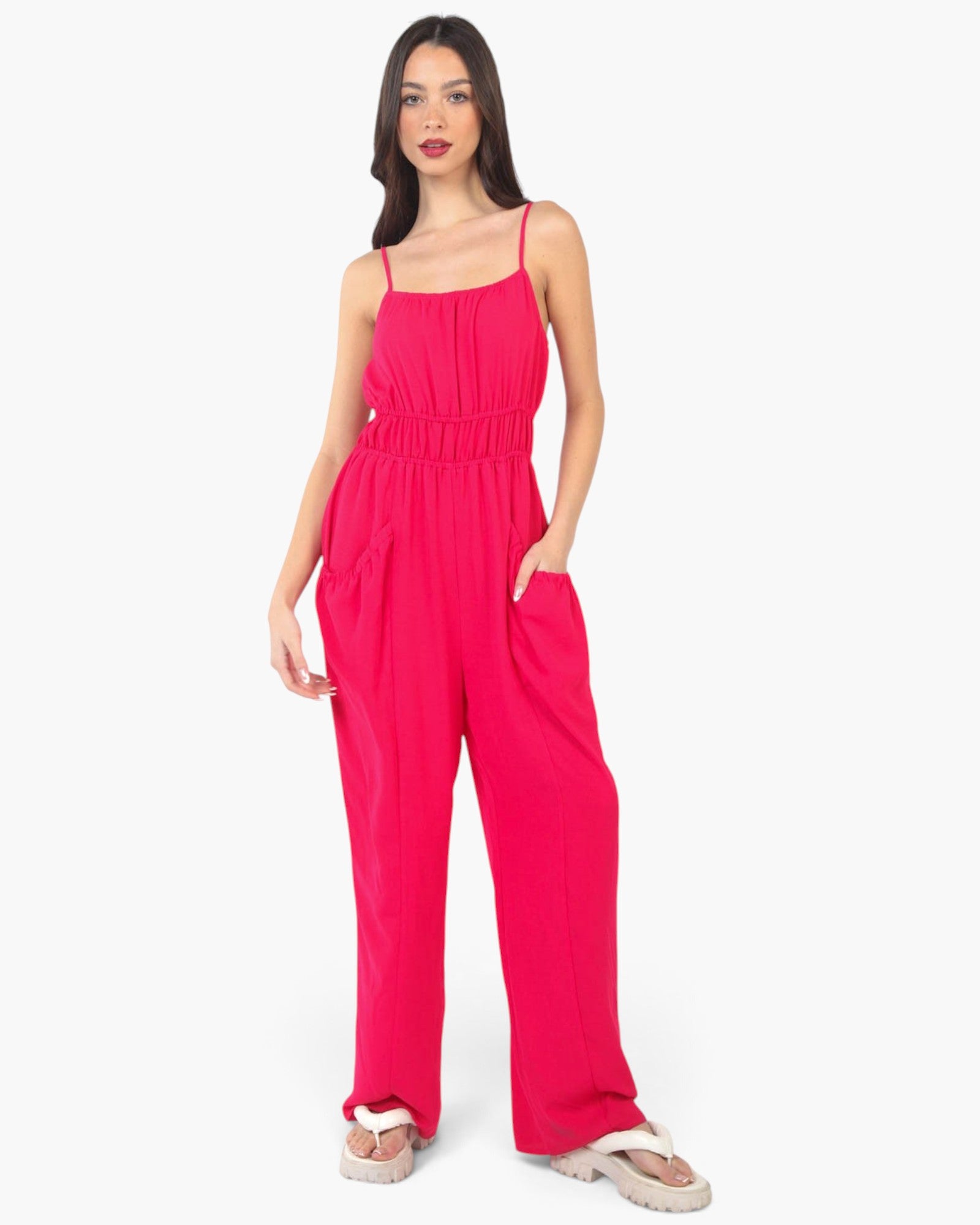 A woman modeling a bright pink sleeveless jumpsuit with thin spaghetti straps, a gathered smocked bodice, and wide legs, paired with white slide sandals against a plain white studio background.