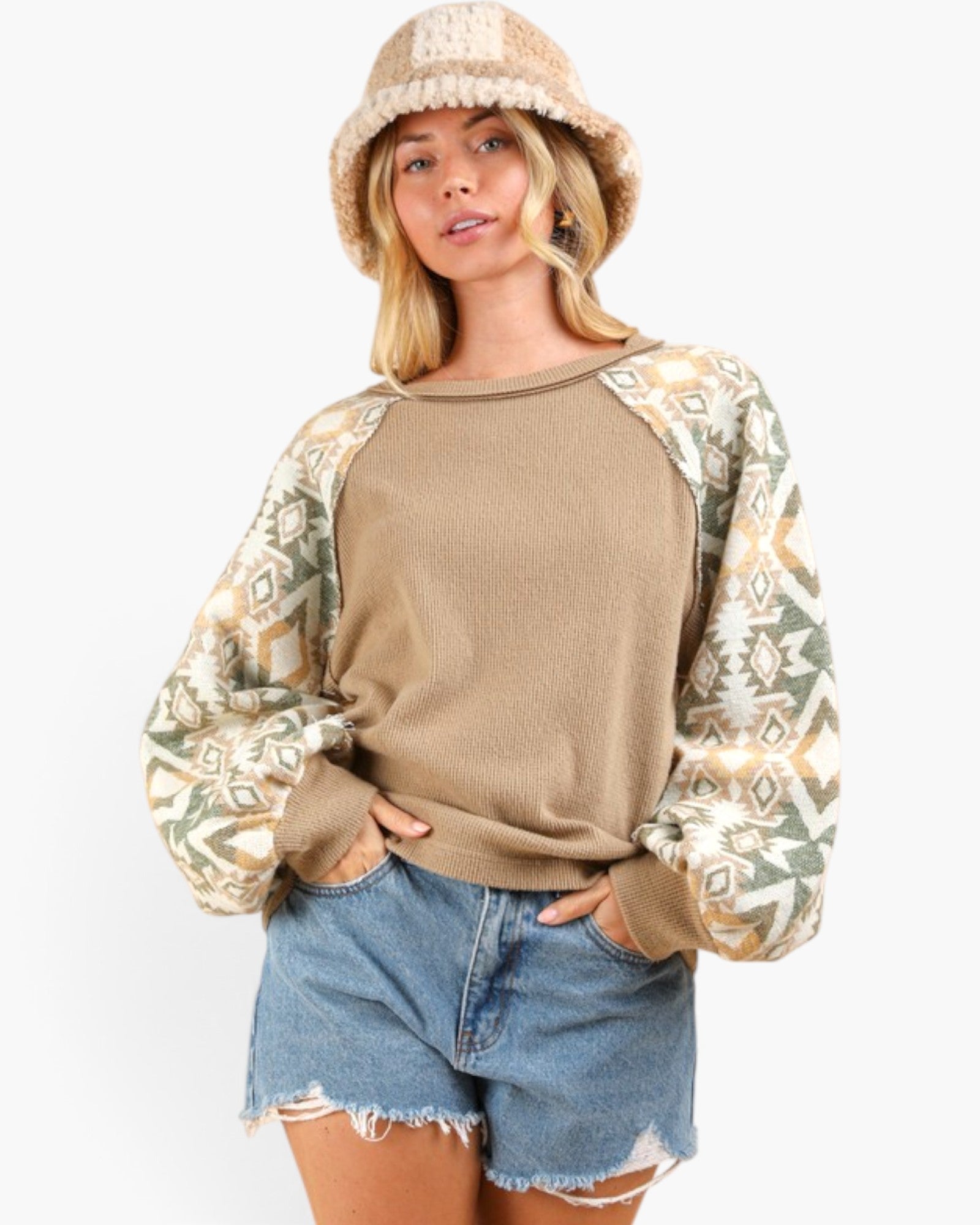 A woman wearing a taupe knit raglan sweatshirt with patterned sleeves, paired with denim shorts and a knit bucket hat, against a plain white background.