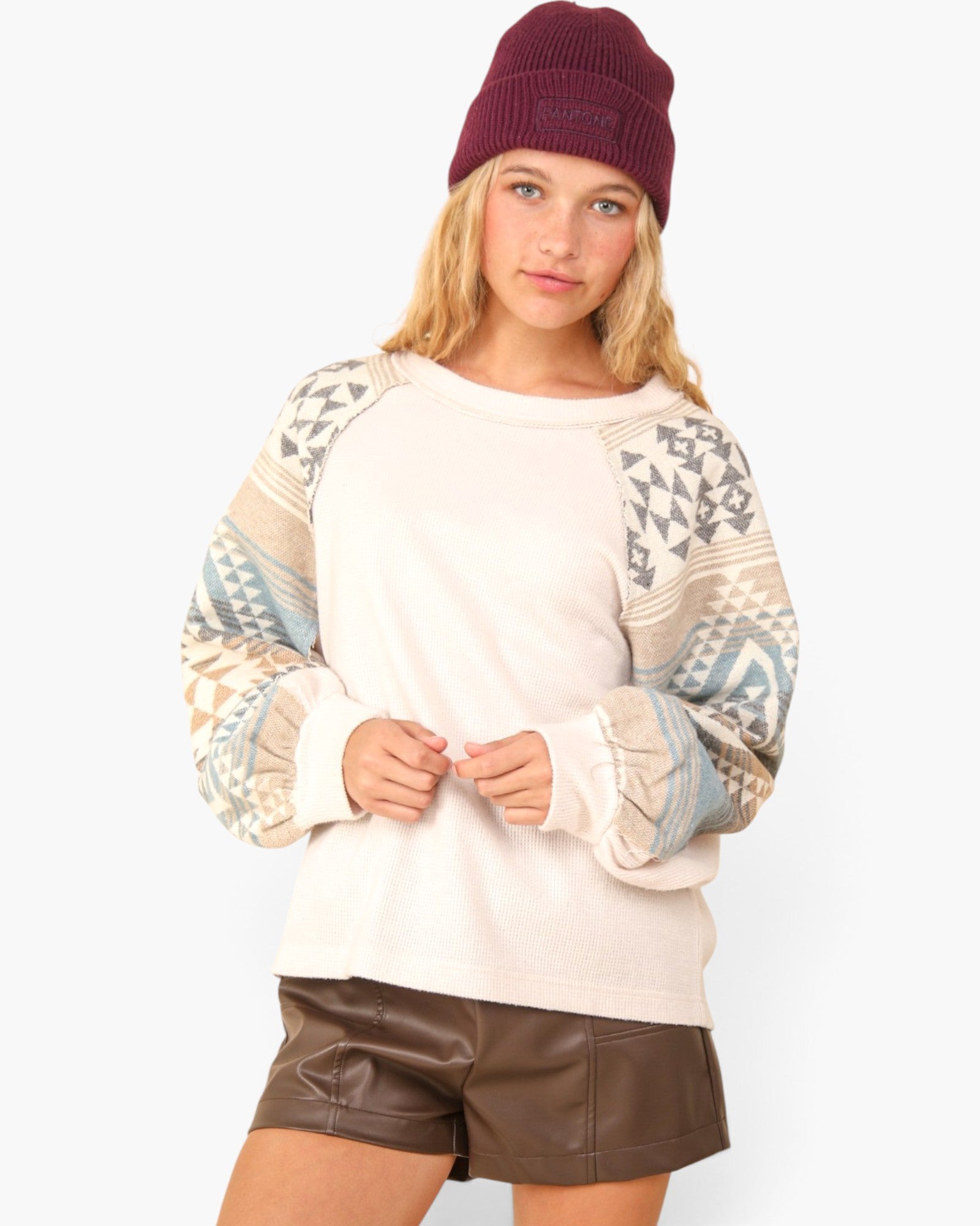 A woman wearing a cream-colored long-sleeve sweatshirt with geometric patterned raglan sleeves, paired with brown faux-leather shorts and a burgundy knit beanie, against a light gray studio background.