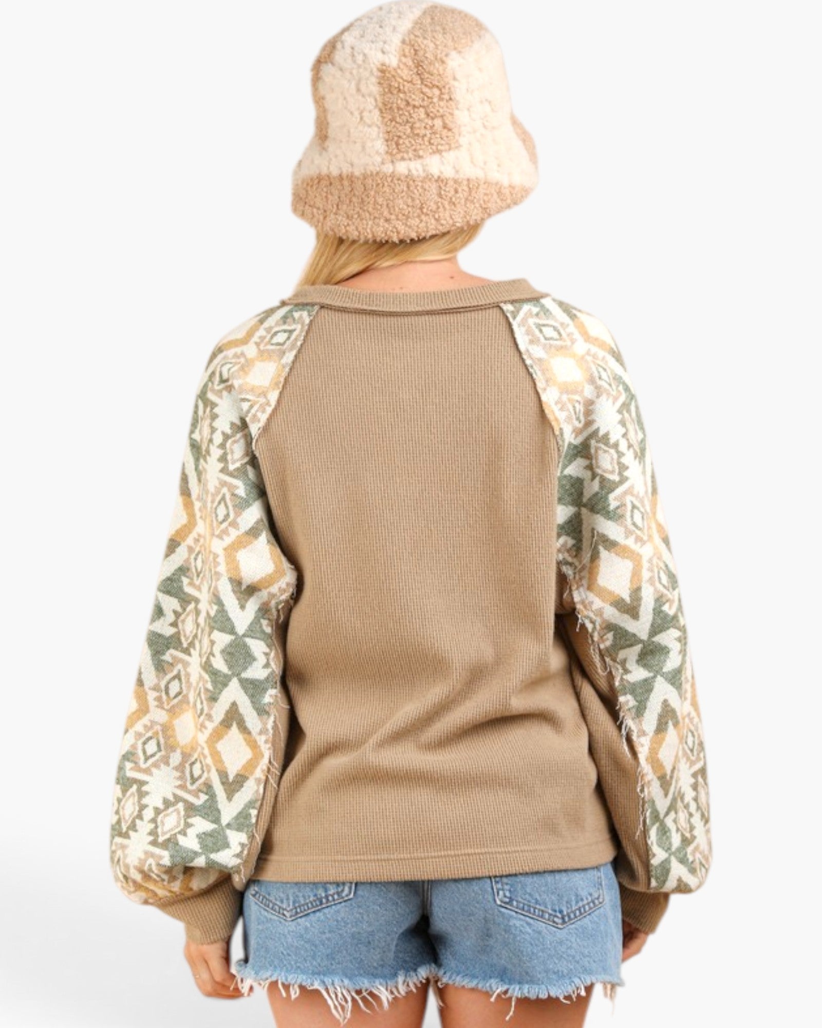A person shown from the back wearing a tan knit sweater with patterned raglan sleeves, a fuzzy beige and cream beanie, and denim shorts against a plain light background.