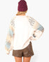 A person shown from the back wearing a cream-colored long-sleeve sweatshirt with geometric patterned raglan sleeves, paired with brown shorts and a burgundy beanie, against a plain light gray background.