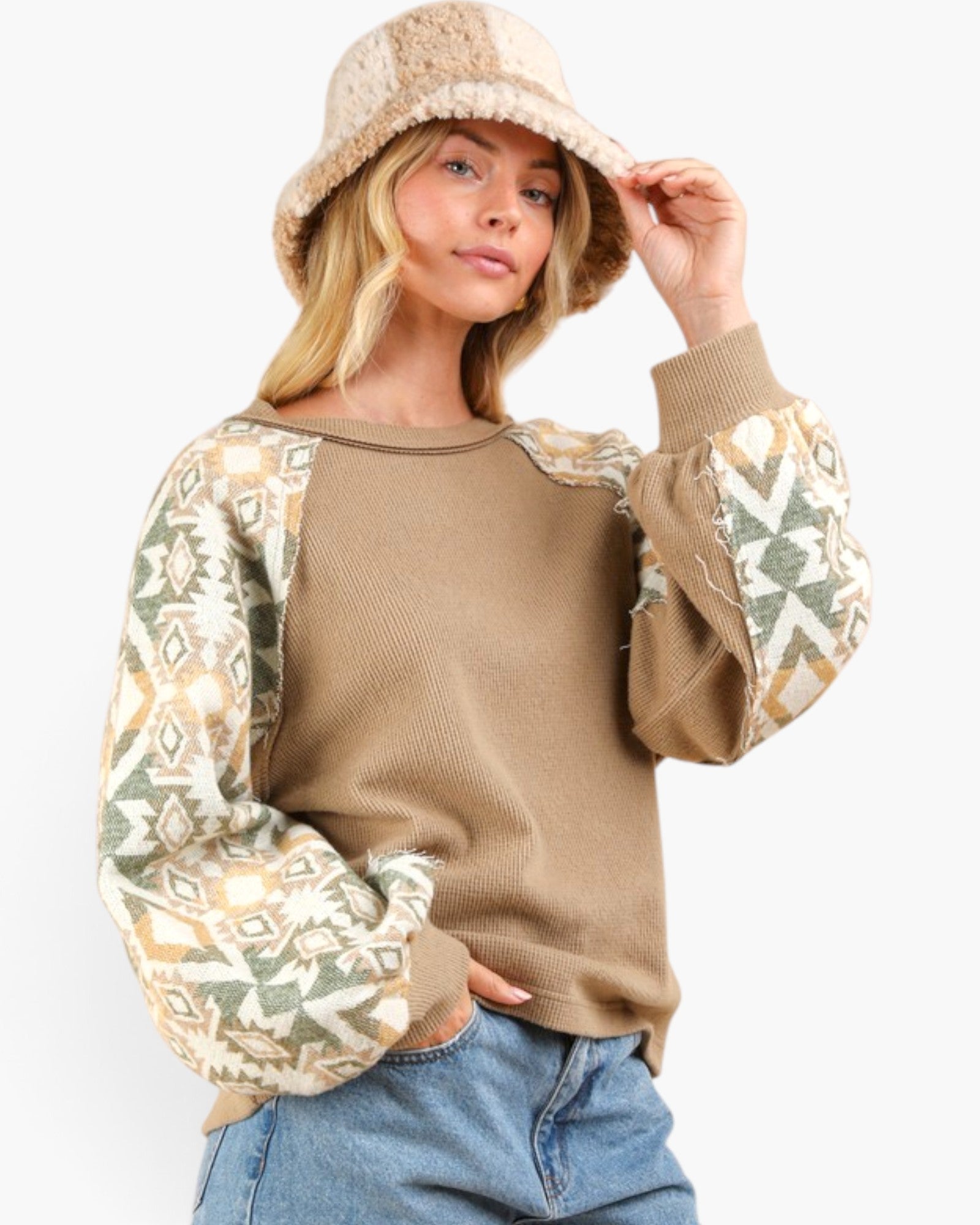 A woman wearing a tan knit sweater with patterned sleeves, a beige bucket hat, and blue jeans, photographed against a plain light gray studio background.