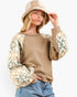 A woman wearing a tan knit sweater with patterned sleeves, a beige bucket hat, and blue jeans, photographed against a plain light gray studio background.