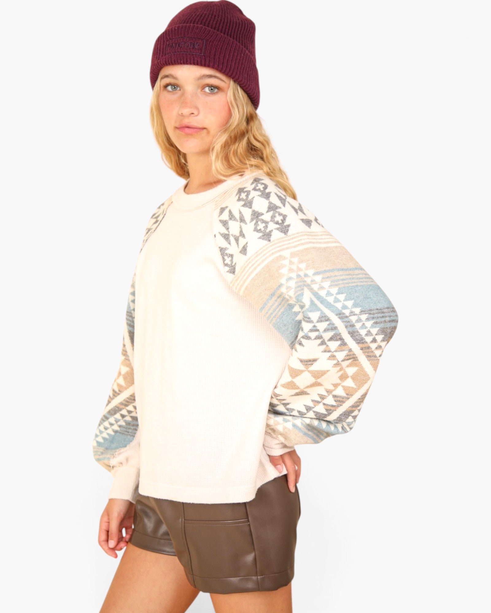 A woman wearing a cream-colored knit sweater with a geometric patterned design on the sleeves, styled with a burgundy beanie and brown shorts against a light gray background.