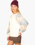 A woman wearing a cream-colored knit sweater with a geometric patterned design on the sleeves, styled with a burgundy beanie and brown shorts against a light gray background.