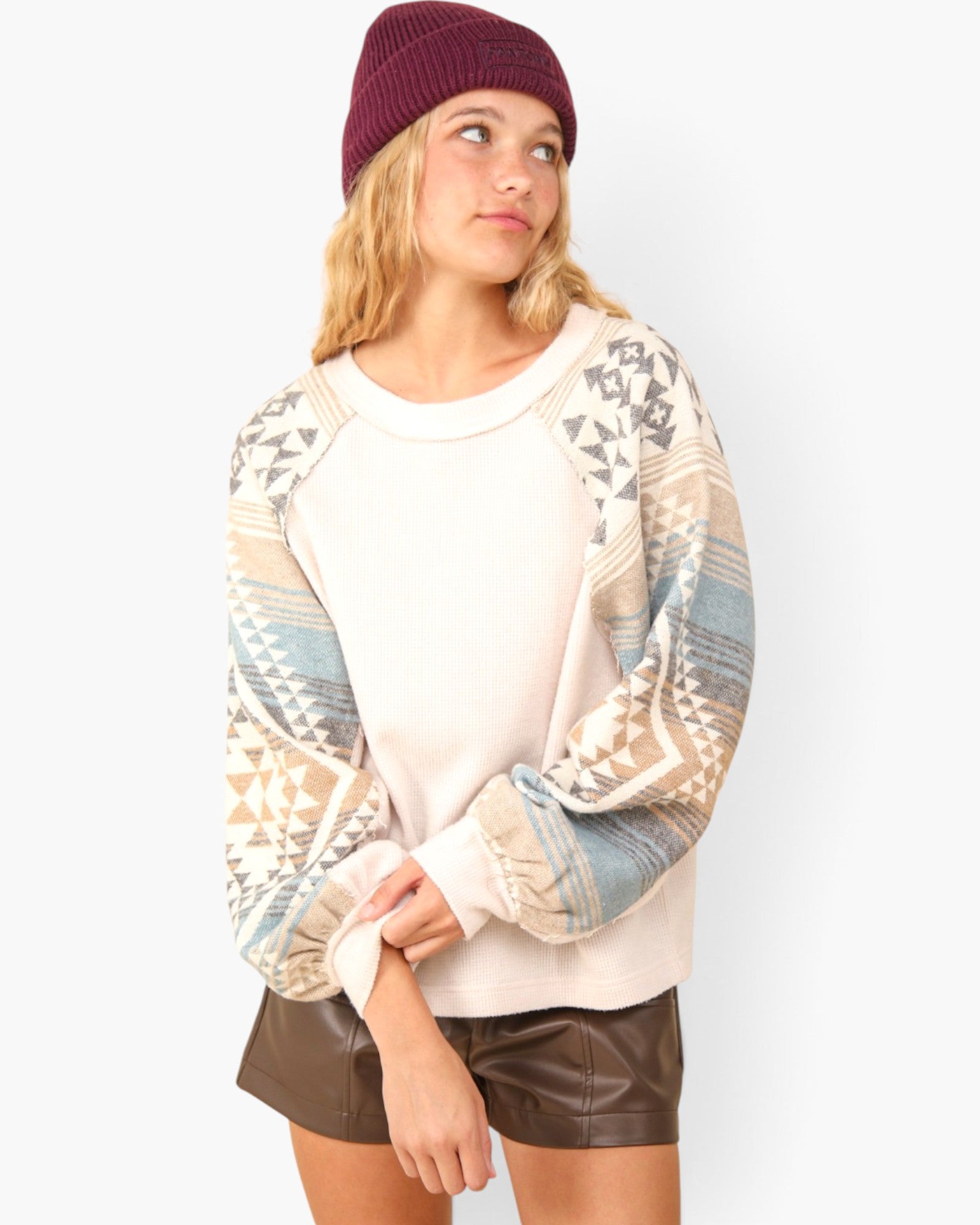 A woman wearing a cream-colored crewneck sweatshirt with patterned raglan sleeves in beige, blue, and gray, paired with brown faux-leather shorts and a burgundy beanie, against a plain white background.