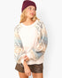 A woman wearing a cream-colored crewneck sweatshirt with patterned raglan sleeves in beige, blue, and gray, paired with brown faux-leather shorts and a burgundy beanie, against a plain white background.