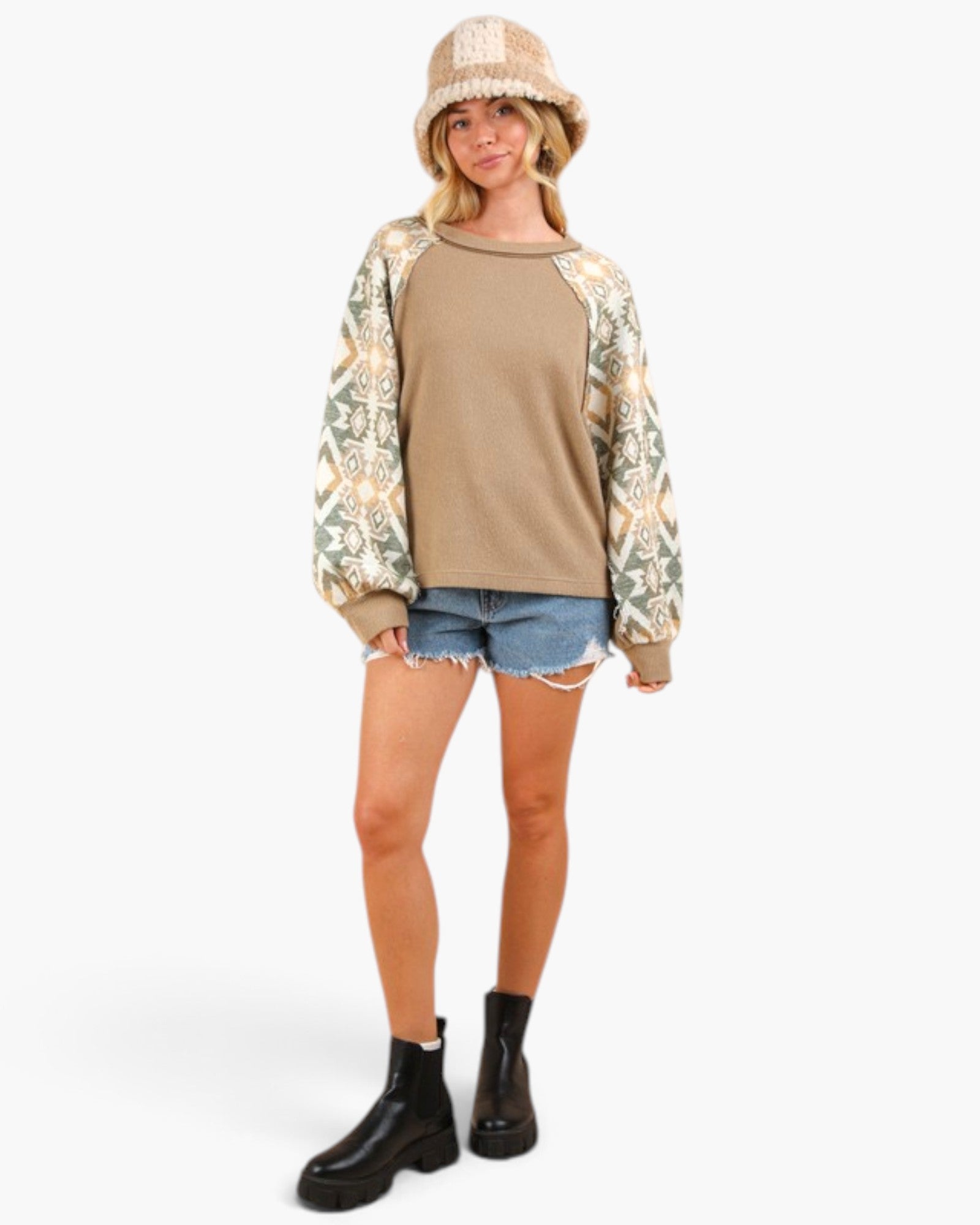 A woman modeling a long-sleeve raglan sweatshirt with a solid tan body and patterned sleeves in beige, green, and cream, paired with denim shorts, a knit bucket hat, and black boots against a light gray background.