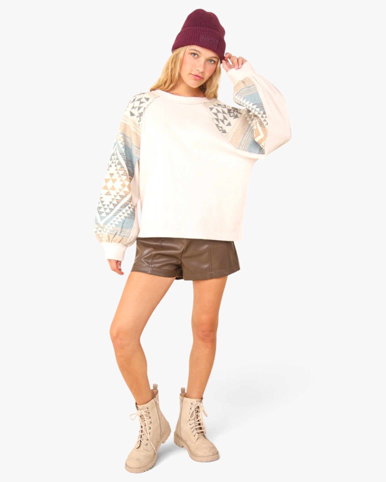 A woman modeling a cream-colored crewneck sweatshirt with patterned sleeves, paired with brown shorts, beige combat boots, and a burgundy beanie, against a light gray background.