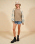 Person wearing a patterned jacket, beige top, denim shorts, and black boots on a beige background
