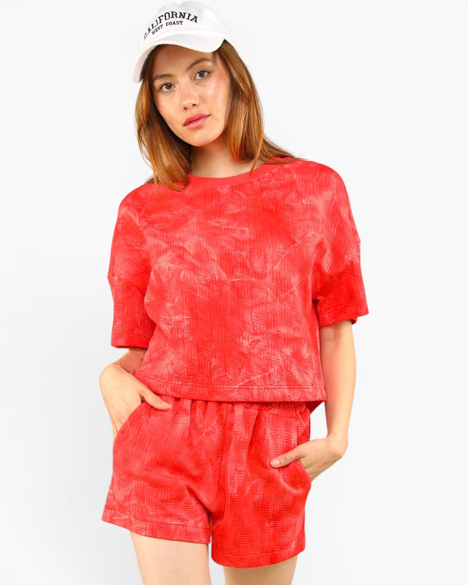 A woman modeling a matching red textured two-piece set consisting of a short-sleeve cropped top and high-waisted shorts, paired with a white baseball cap.