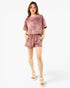 A woman modeling a matching two-piece lounge set consisting of a short-sleeve t-shirt and shorts in a dusty rose pink color with a subtle marbled texture, paired with white platform sandals, against a plain white studio background.