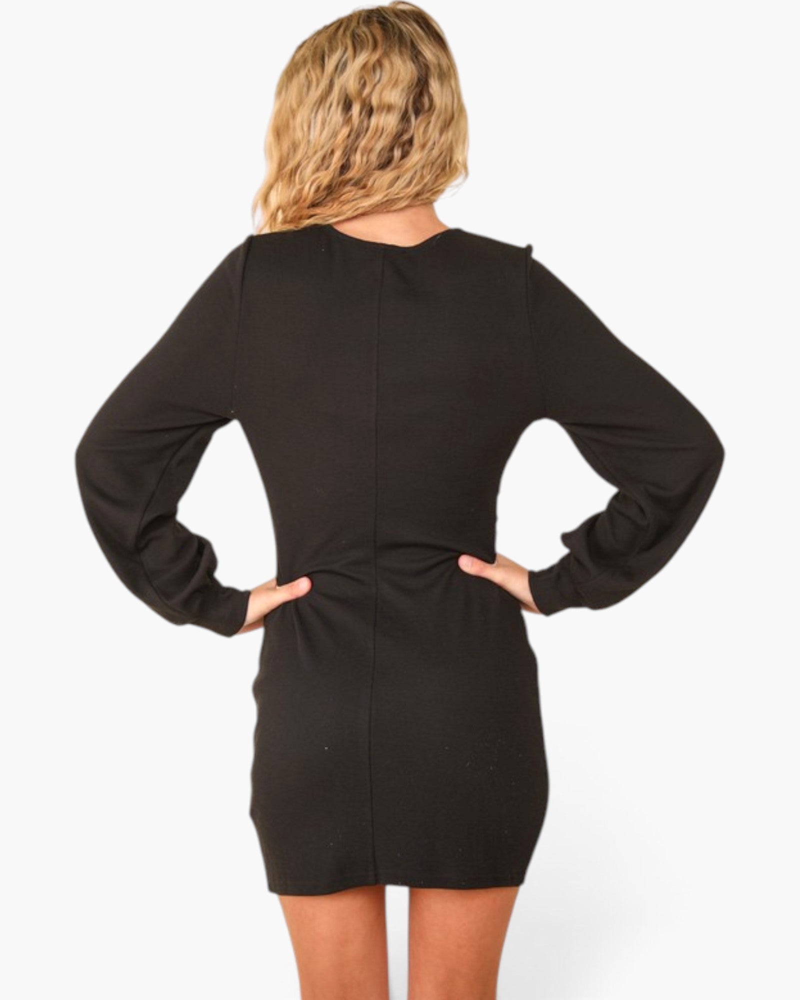 A woman shown from the back wearing a black long-sleeve mini dress, standing with hands on hips against a plain white studio background.
