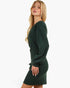 A blonde model wearing a dark forest green long-sleeve mini dress with a tie detail at the waist, shown in profile against a plain white background.