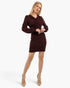 A blonde model wearing a dark burgundy long-sleeve mini dress with a wrap-style V-neckline, styled with beige lace-up ankle-strap heels against a light gray studio background.